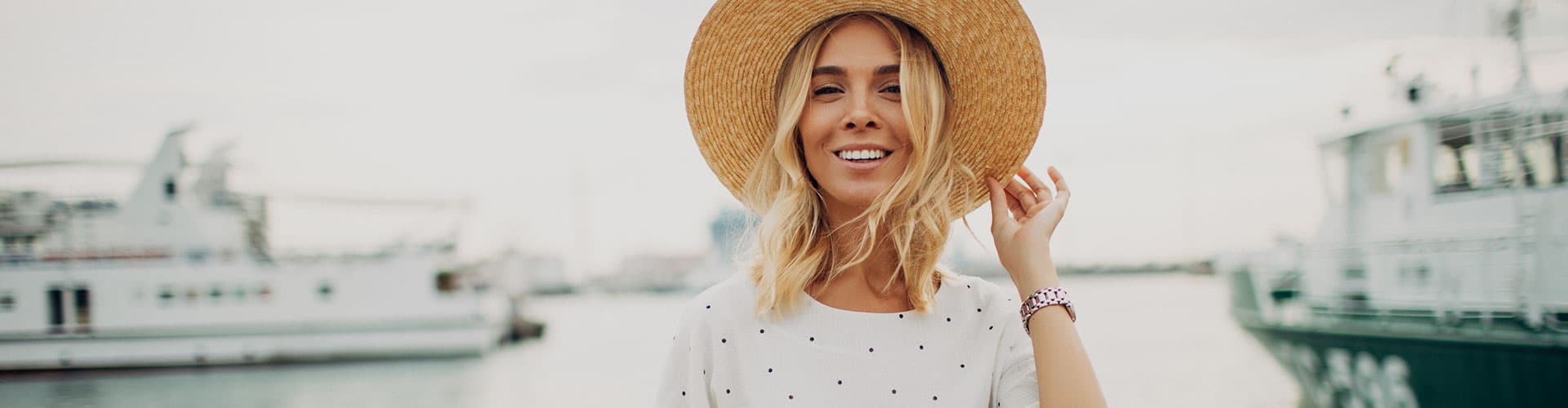 A smiling woman with blonde hair wearing a wide-brimmed straw hat and a white polka-dot shirt stands near a harbor with boats in the background. Bella Lei Aesthetics and Wellness - Flemming Island, Florida