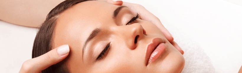 A woman with closed eyes receives a gentle facial massage, with hands gently touching her temples, promoting relaxation and calmness. Bella Lei Aesthetics and Wellness - Flemming Island, Florida