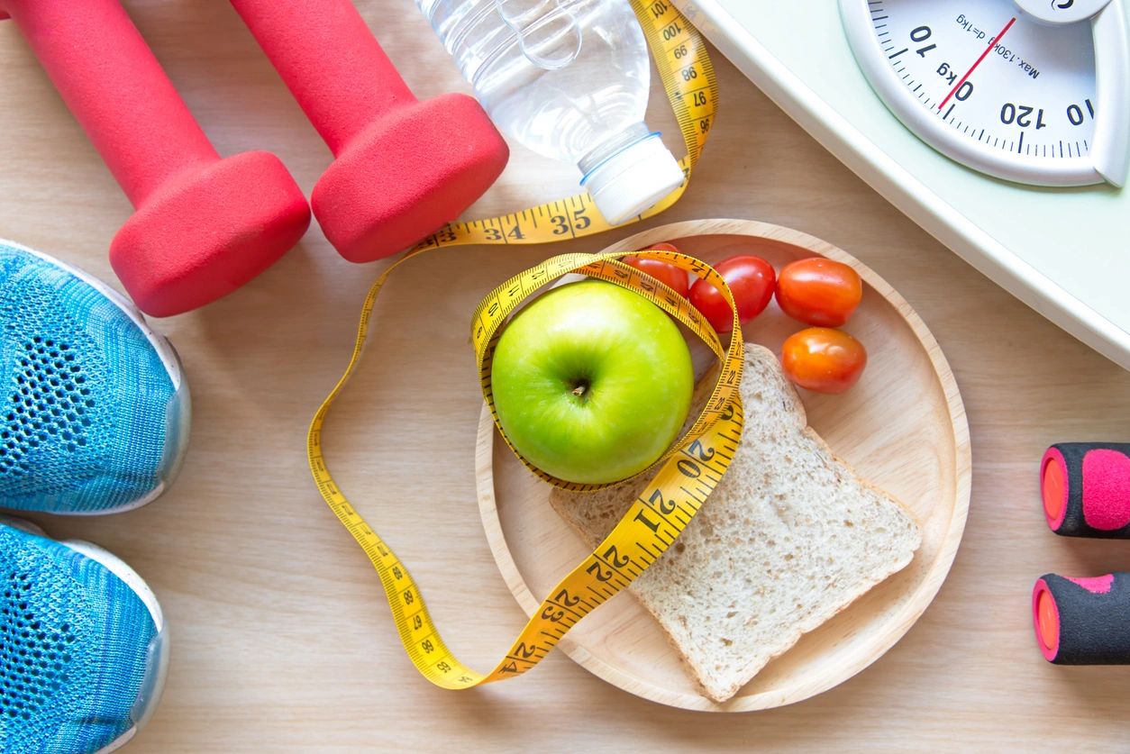 A plate with an apple, grape tomatoes, and whole grain bread is surrounded by a measuring tape, water bottle, scale, red dumbbells, workout gloves, and blue sneakers, symbolizing a healthy lifestyle. Bella Lei Aesthetics and Wellness - Flemming Island, Florida