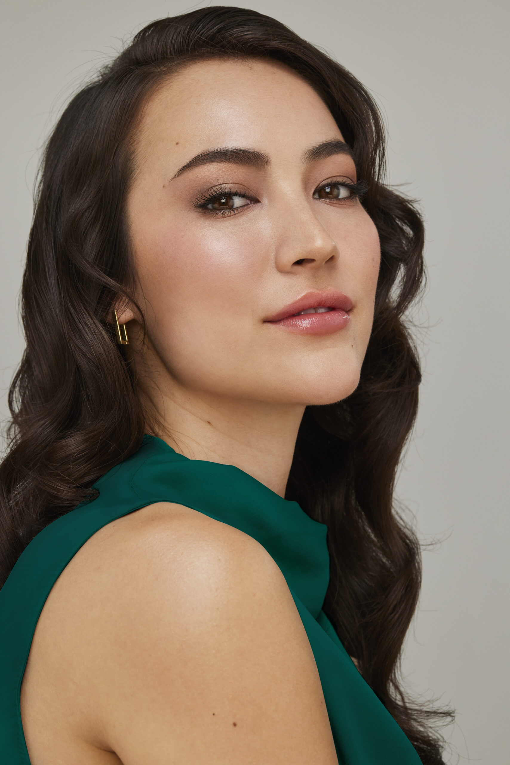 A woman with long, wavy dark hair and glowing skin looks confidently at the camera. She wears a sleeveless green top, subtle makeup, and a small gold earring, against a plain light background. Bella Lei Aesthetics and Wellness - Flemming Island, Florida